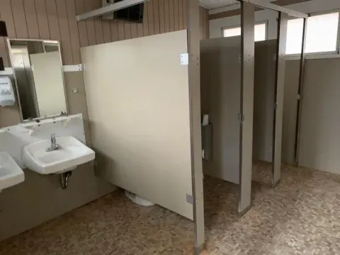 Partial interior view of comfort station. 2 small bathroom stalls, 2 windows, 2 porcelain sinks with mirrors. Flooring is tile,
