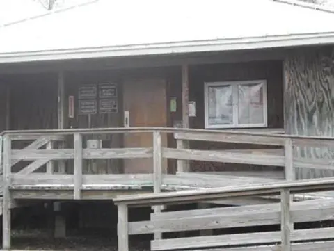 Wooden building entrance with a ramp and railings leading to a covered porch, a closed door, posted signs, and a window displaying notices.