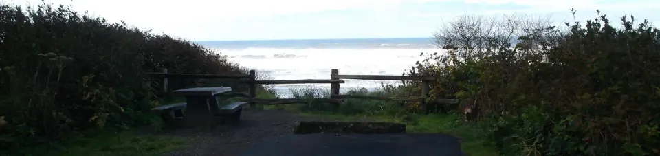 A campsite on a bluff overlooking the Pacific Ocean. The campsite has a paved parking spot as well as a picnic table and fire ring.