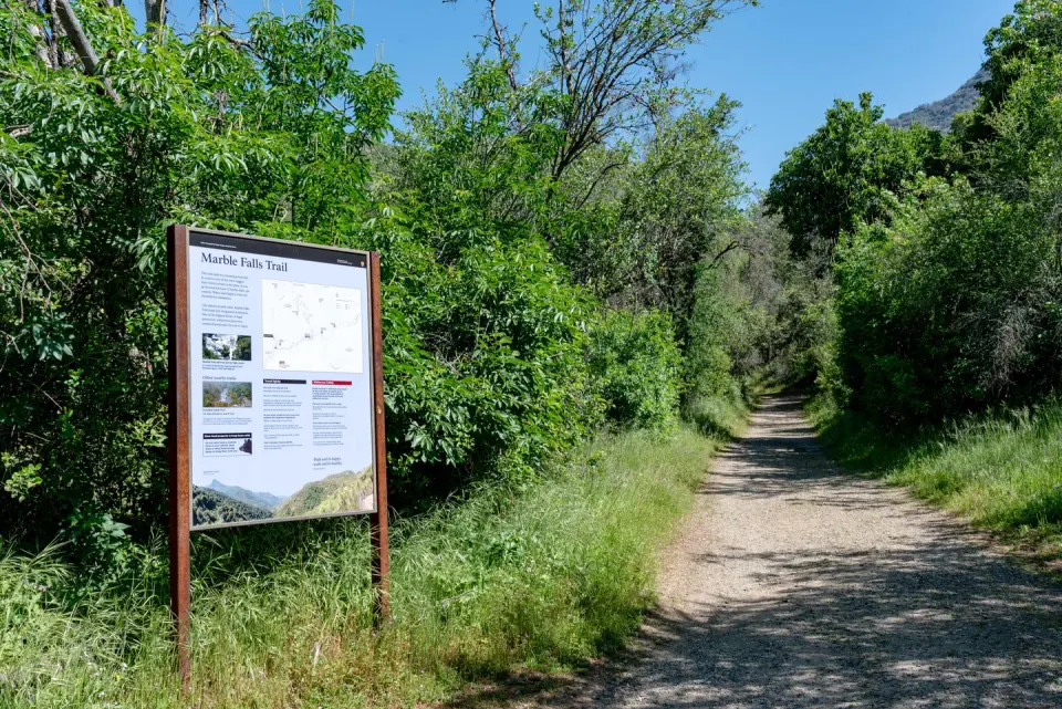 Trailhead and sign for the marble falls trail. On the left is the trail sign, with information about the trail and what you will find on it. To the right is the trail itself, dirt and gravel, with grass and local trees growing up alongside it. It is very green because this is an image from spring. It is not normally green.