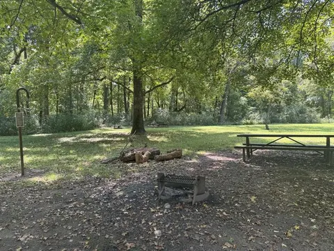 A brown picnic table sits to the right of a campsite. A campfire ring and lantern post can be seen in the foreground.