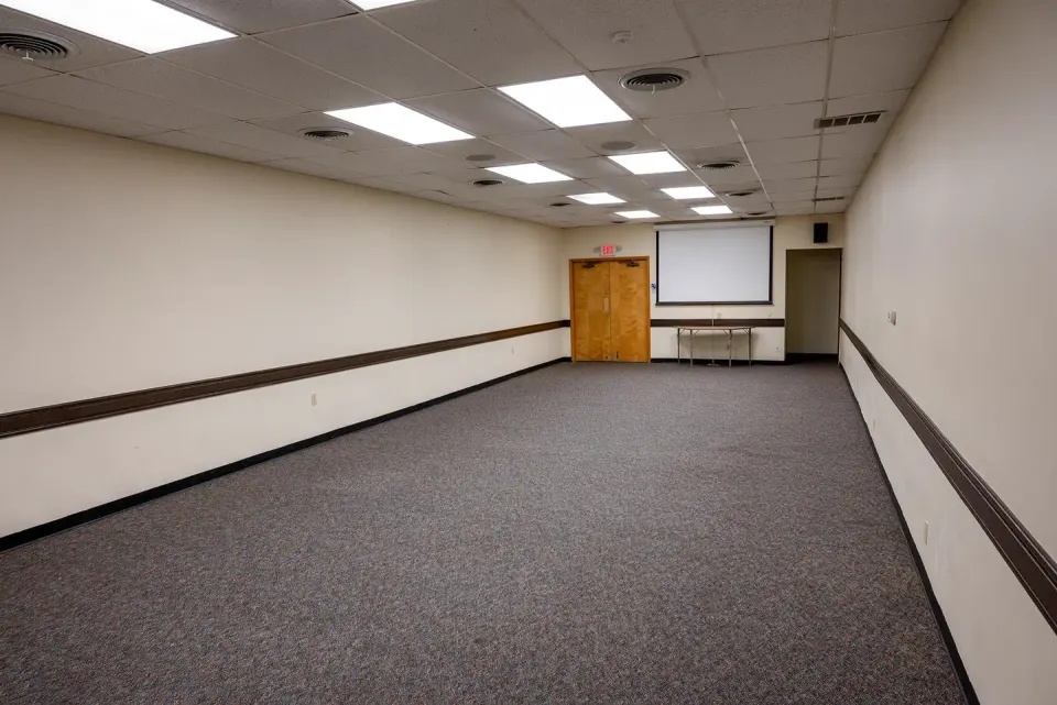 Interior view of the small conference room. Gray carpet flooring with 2 doors at the far end. Ceiling mounted video screen on far end of room.