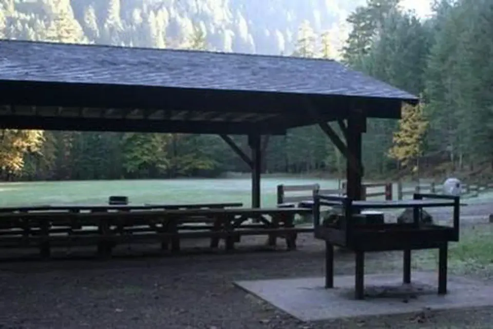 Group picnic shelter and barbecue next to large flat lawn ringed by mostly conifer forest.