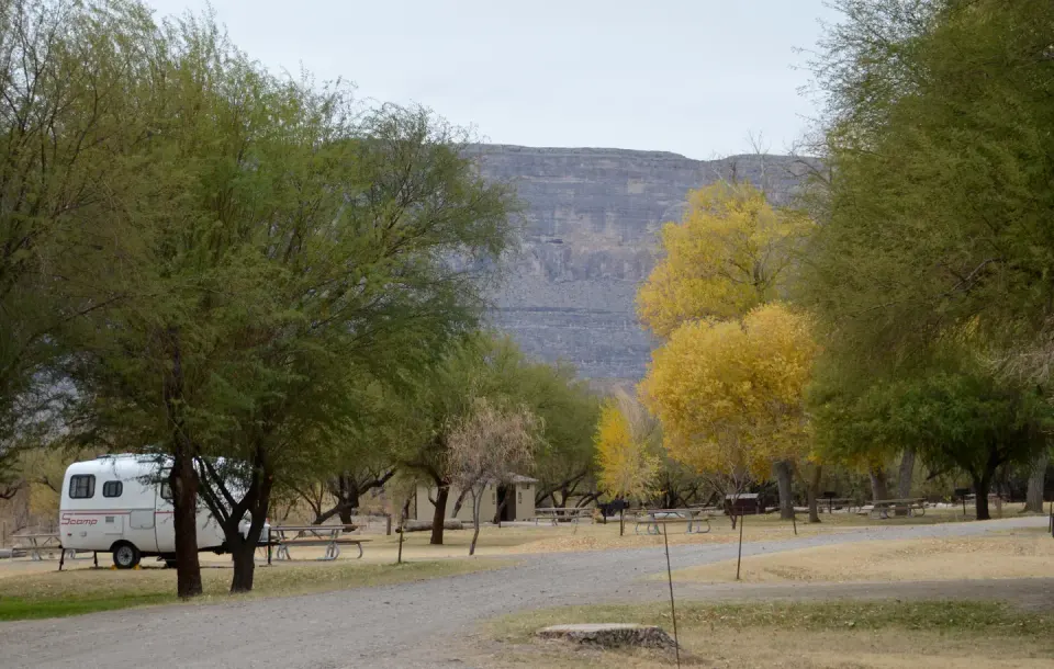 Small travel trailer set up in tree-lined campground with cliffs in background
