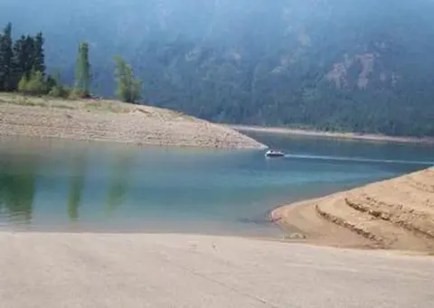 View of boat ramp leading into lake with sloped earthen banks, looking beyond to a small boat and tree covered slopes above high water line.