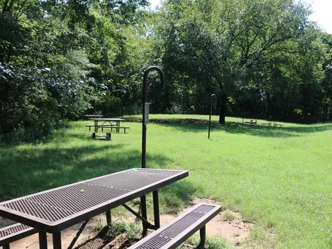 A picnic table sits on a green lawn with other picnic tables and trees in the background.