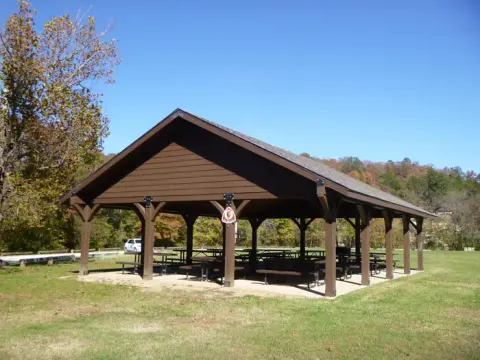 Front view of the covered pavilion with picnic tables surrounded by a grassy field. Green trees beginning to change colors are visible in the bakckground.