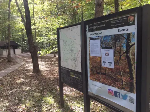 Bulletin board and map sign in the forest near a sidewalk and building in a campground