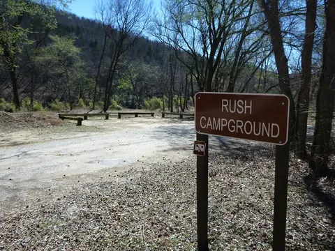 A brown sign stating "Rush Campground" can be seen in the foreground with a dirt road, trees, and a parking area in the background.