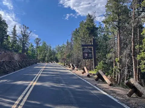 The road leading up to Longs Peak Campground and the Longs Peak Ranger Station. Wooden parking barriers line the road, and there is a location sign on the right.
