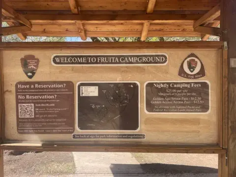A Bulletin Board with 7 signs in it. The signs are as follows, starting at the top center and moving clockwise: "Welcome to Fruit Campground"; "U.S. Fee Area"; "Night Camping Fees. $25.00 per site; Maximum of 8 people per site. Golden Age/Senior Pass- $12.50; Golden Access/Access Pass- $12.50. No discount with National Parks and Federal Recreation Lands Annual Pass"; Map of the campground; "See back of sign for park information and regulations"; "Have a Reservation? Refer to the campground map and proceed to your campsite. Don't know your campsite number? Go to the visitor center to connect to free public Wi-Fi to access your reservation confirmation. Wi-Fi is available 24/7. No Reservation? All sites must be reserved through Recreation.gov. Go to the visitor center to connect to free public Wi-Fi to reserve a site. Wi-Fi is available 24/7. Visitor center staff cannot reserve the site for you. The top right corner of the same sign reads as follows: "Seasons. Loops A & B: Open year-round. Loop C: Open March 1st- End of November."; The NPS Arrowhead.