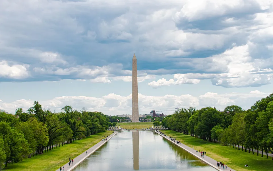 A scenic view of the Washington Monument in Washington, D.C., with reflections in the reflecting pool and a cloudy sky overhead.
