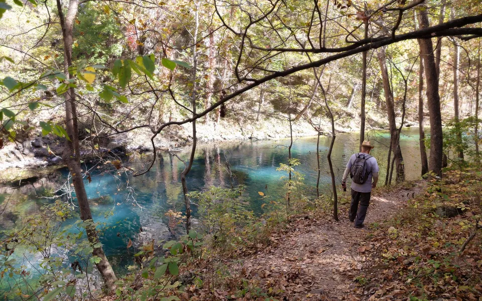 A hiker walks along a scenic trail beside a vibrant blue river in Camdenton, Missouri during the colorful fall season.