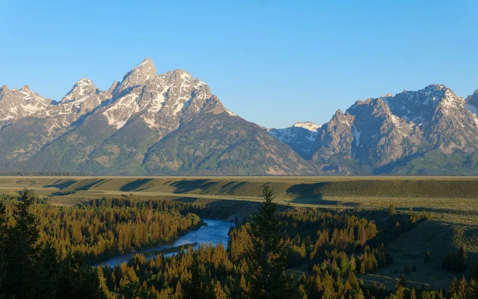 Experience the stunning Grand Teton mountains with vibrant greenery and a serene river at sunrise in Wyoming.